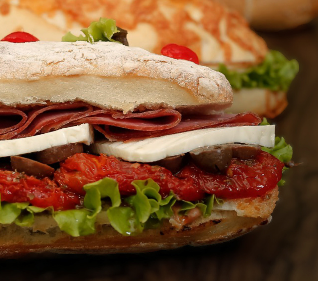 Foto de sanduíche de metro recheado com salame, alface, tomate e decorado com cereja no topo.