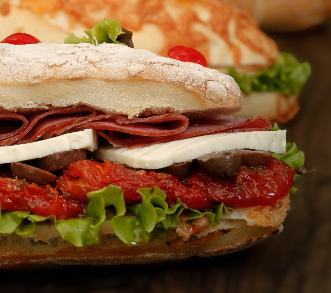Foto de sanduíche de metro recheado com salame, alface, tomate e decorado com cereja no topo.